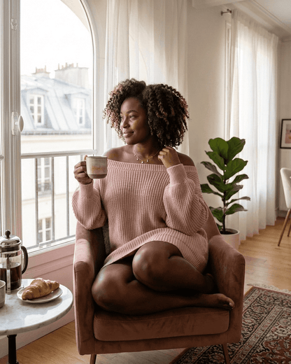 Woman in pink sweater dress sitting in a chair, enjoying a coffee with a cozy vibe.