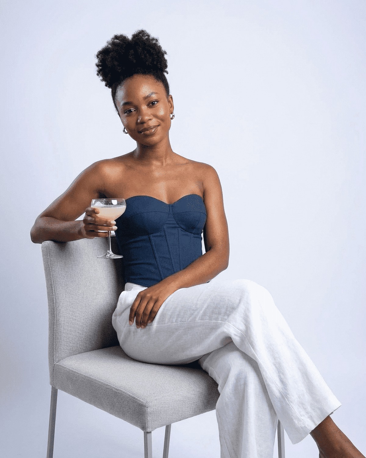 Model wearing the Atlas Corset Top in navy faux denim, holding a cocktail glass while sitting on a chair.