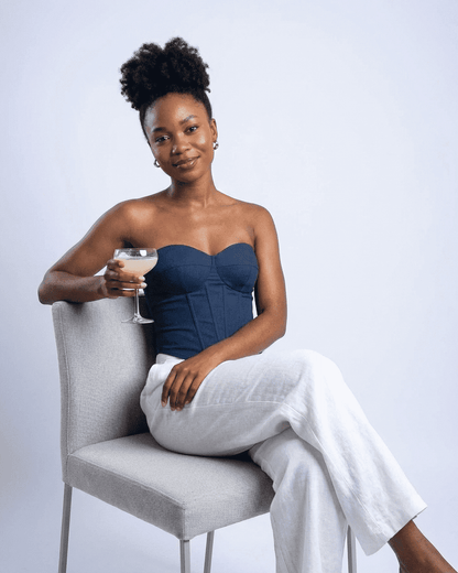 Model wearing the Atlas Corset Top in navy faux denim, holding a cocktail glass while sitting on a chair.