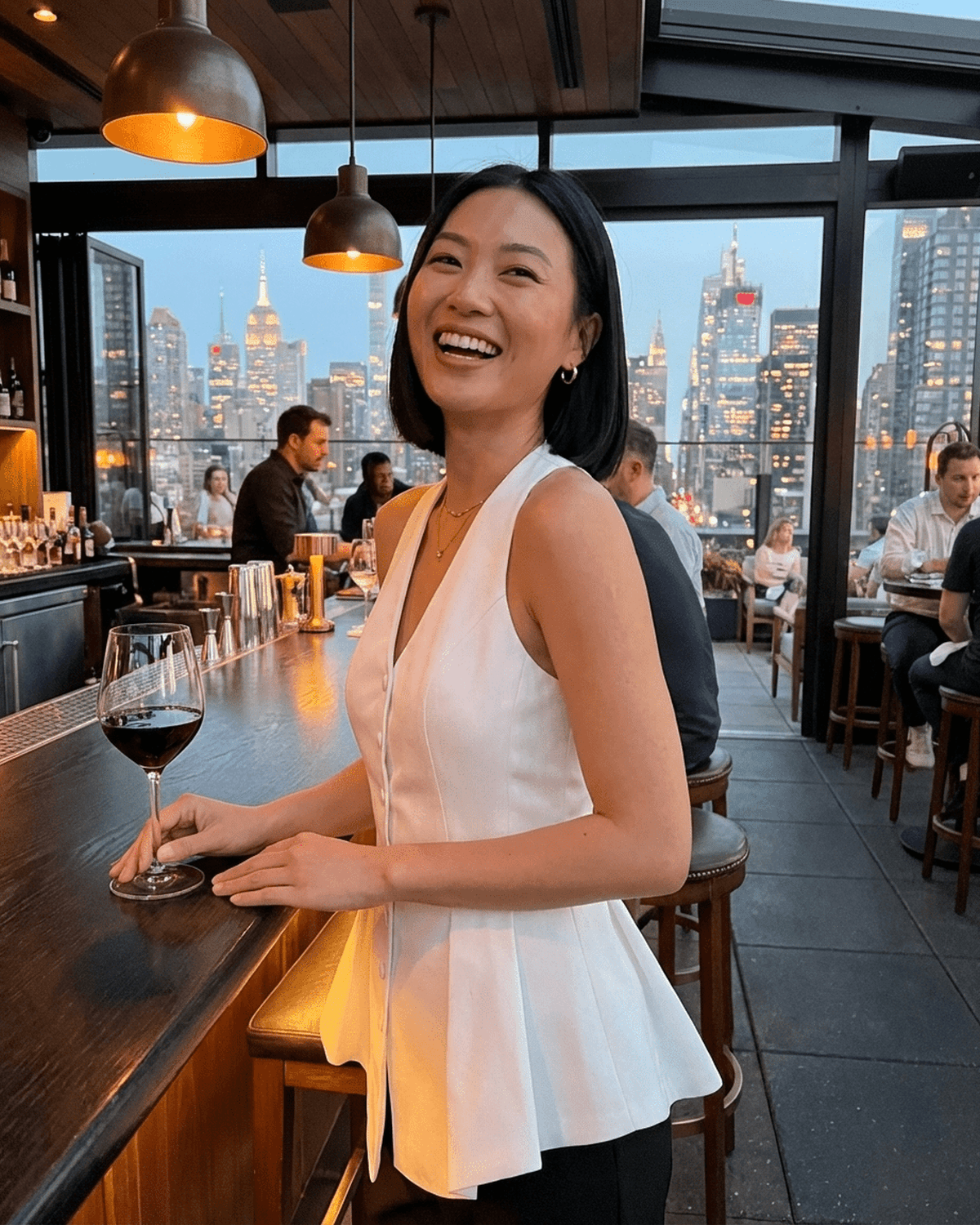 Woman wearing a Celeste Blouse by RAMBLION, smiling at a bar with a wine glass in hand, city skyline in the background.