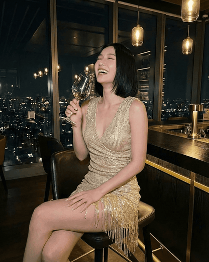 A woman laughing in an Aurora Mini Dress by RAMBLION, featuring gold sequins and fringe hem, holding a glass of wine.