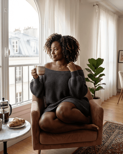 Woman in a cozy off-the-shoulder sweater enjoying coffee at home, showcasing the Ember Mini Dress from RAMBLION.
