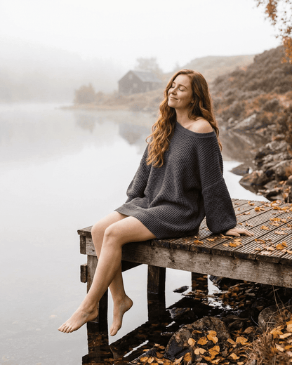 RAMBLION Ember Mini Dress model sitting by a lake, showcasing the cozy charcoal ribbed knit fabric and off-the-shoulder design.