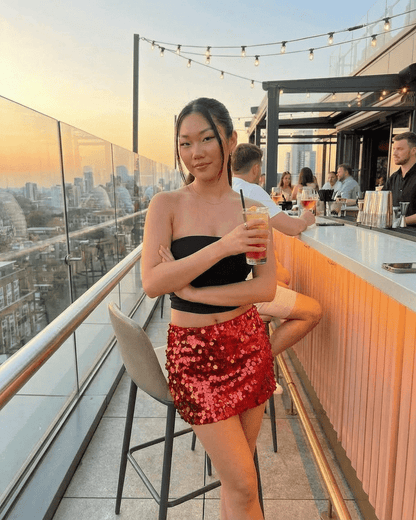 Model wearing RAMBLION Flamenco Mini Skirt with red sequins, enjoying a cocktail at a rooftop bar during sunset.