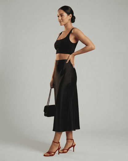 Model wearing RAMBLION Noirelle Midi Skirt, a sleek satin mermaid midi skirt, styled with a black top and red heels.