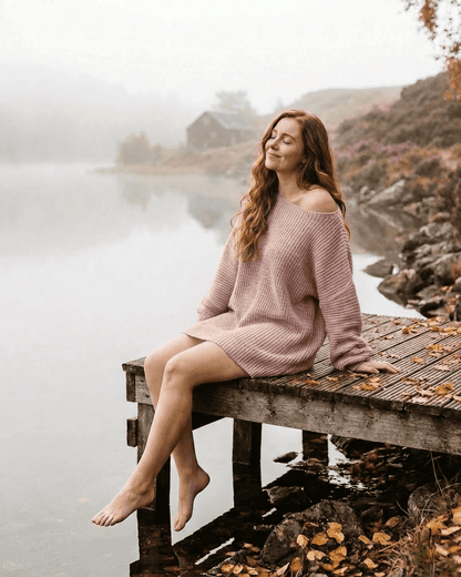 Nova Mini Dress by RAMBLION, showcasing a model in a soft pink ribbed knit off-the-shoulder design by a serene lake.