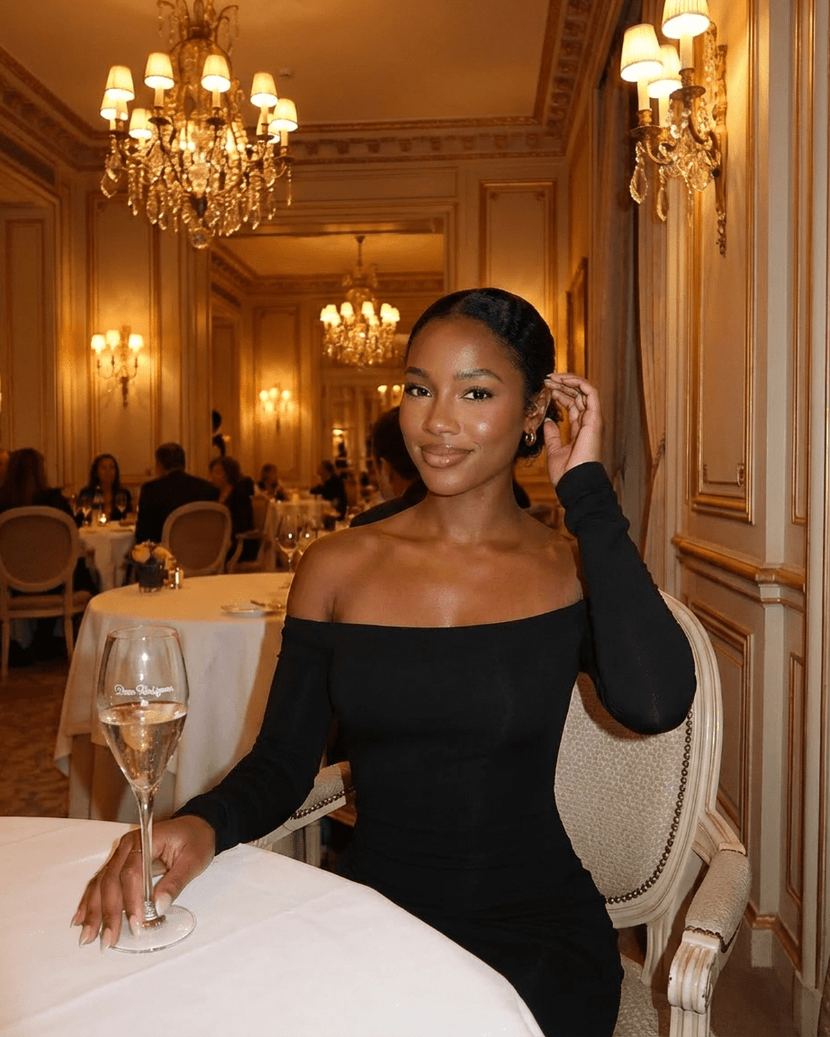 Elegant woman in Nocturne Maxi Dress by RAMBLION, off-shoulder style, seated in a luxurious dining setting.