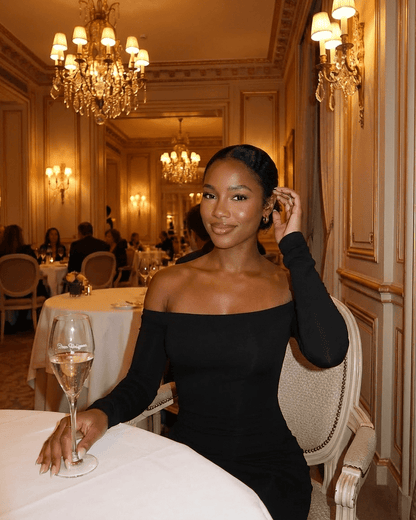 Elegant woman in Nocturne Maxi Dress by RAMBLION, off-shoulder style, seated in a luxurious dining setting.