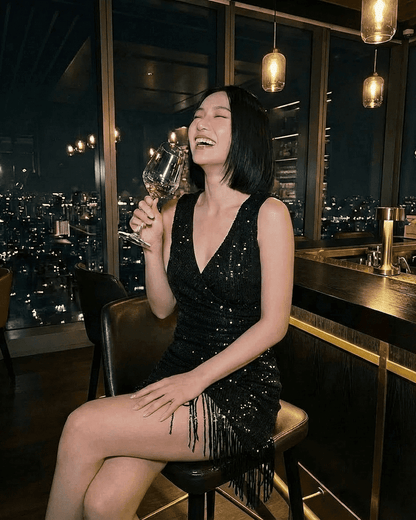 RAMBLION Noir Mini Dress worn by a woman laughing while holding a wine glass, featuring a black sequin design and fringe hem.