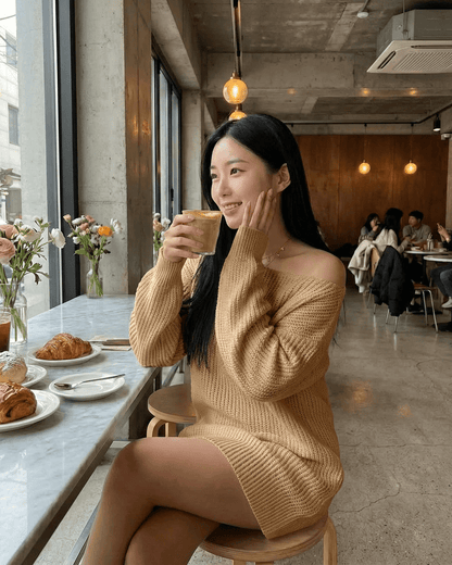 Woman enjoying a coffee in a cozy café wearing a stylish off-the-shoulder sweater dress, natural light and floral decor.