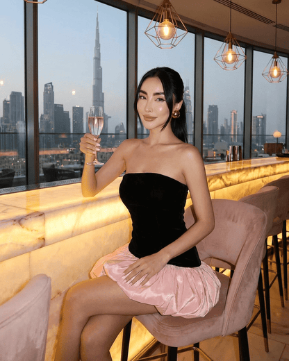 Selina Mini Dress model featuring black velvet strapless top and pink puff skirt in a stylish bar with skyline view.